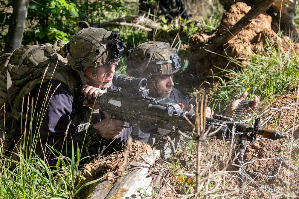 Two soldiers in full combat gear are positioned in a prone firing position behind a dirt berm in a wooded area. One soldier is actively aiming a belt-fed machine gun equipped with an optical scope, while the second soldier observes alongside. Both wear modern tactical helmets with night vision device mounts and camouflage uniforms, suggesting a military training exercise or field operation.