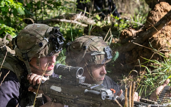 Two soldiers in full combat gear are positioned in a prone firing position behind a dirt berm in a wooded area. One soldier is actively aiming a belt-fed machine gun equipped with an optical scope, while the second soldier observes alongside. Both wear modern tactical helmets with night vision device mounts and camouflage uniforms, suggesting a military training exercise or field operation.