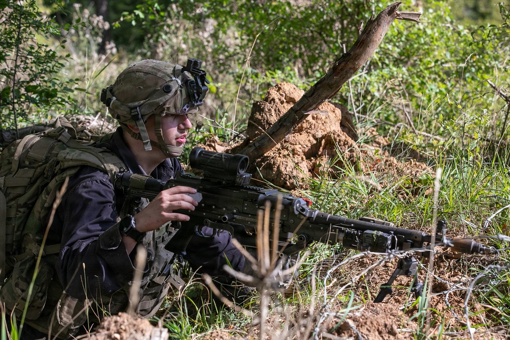 A soldier in full tactical gear is positioned in a prone/low crouch position in a wooded, earthy terrain, aiming a light machine gun mounted on a bipod. The soldier is equipped with a combat helmet fitted with a night vision device mount, tactical vest, backpack, and optical scope on the weapon. The setting appears to be a field training exercise or operational deployment in a forested area.