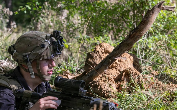 A soldier in full tactical gear is positioned in a prone/low crouch position in a wooded, earthy terrain, aiming a light machine gun mounted on a bipod. The soldier is equipped with a combat helmet fitted with a night vision device mount, tactical vest, backpack, and optical scope on the weapon. The setting appears to be a field training exercise or operational deployment in a forested area.
