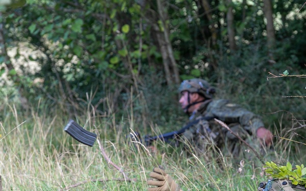 Two soldiers in camouflage uniforms are positioned low in a grassy, wooded area, appearing to conduct a tactical field exercise. The foreground soldier is lying prone and tossing or catching a rifle magazine in the air, while a second soldier is positioned further back in a low crouch. Both wear helmets with camouflage covers and full combat gear.