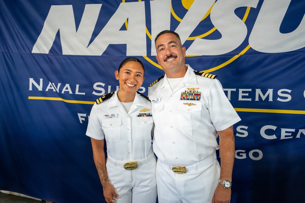 Two U.S. Navy officers in white dress uniforms pose together in front of a Naval Supply Systems Command (NAVSUP) banner, likely at a ceremony or official event in San Diego. The officer on the right displays a significantly higher number of ribbons and insignia, suggesting senior rank. Both are smiling and appear to be posing for an official or commemorative photograph.