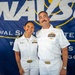 Two U.S. Navy officers in white dress uniforms pose together in front of a Naval Supply Systems Command (NAVSUP) banner, likely at a ceremony or official event in San Diego. The officer on the right displays a significantly higher number of ribbons and insignia, suggesting senior rank. Both are smiling and appear to be posing for an official or commemorative photograph.