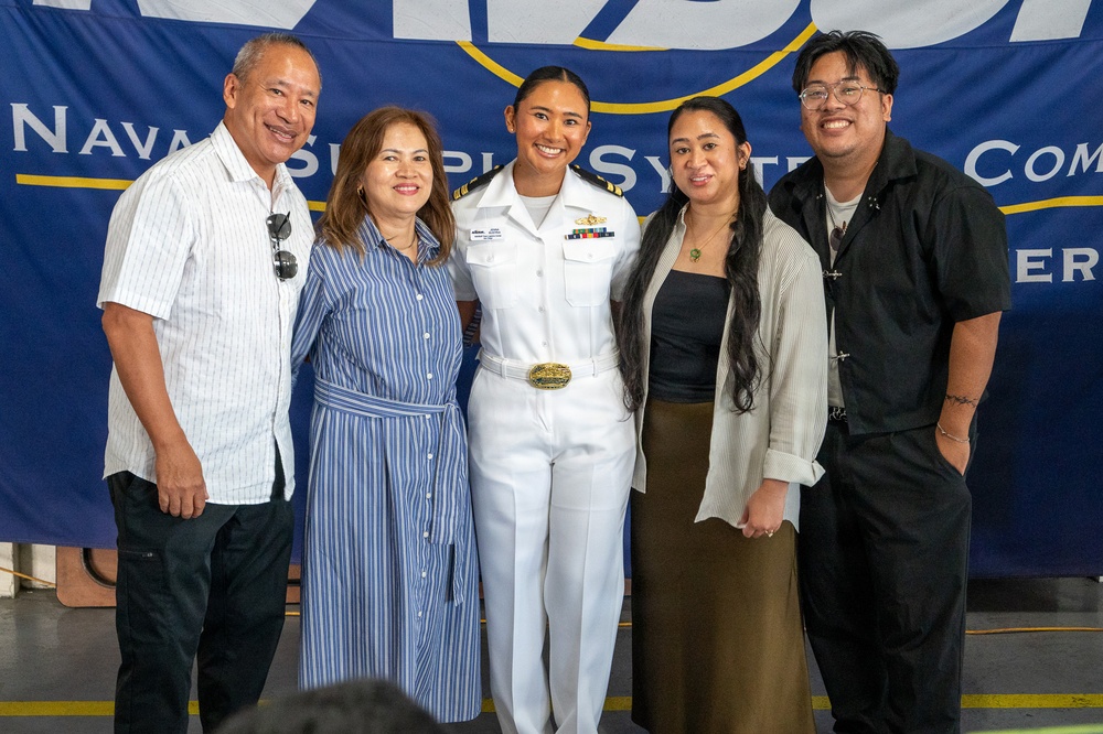 A group of five people pose for a photo in front of a Naval Supply Systems Command banner. The central figure is a female U.S. Navy officer in white dress uniform, surrounded by what appear to be family members. The setting suggests a military ceremony or official event.