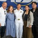 A group of five people pose for a photo in front of a Naval Supply Systems Command banner. The central figure is a female U.S. Navy officer in white dress uniform, surrounded by what appear to be family members. The setting suggests a military ceremony or official event.