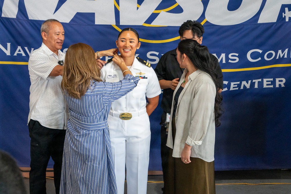 A U.S. Navy officer in white dress uniform is being pinned with new insignia during a promotion or commissioning ceremony. Family members surround her, with one person actively pinning her collar while others look on smiling. The event takes place in front of a Navy banner reading 'Naval... Systems Command... Center.'
