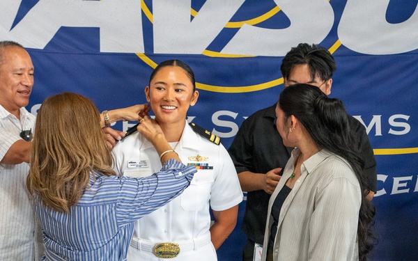 A U.S. Navy officer in white dress uniform is being pinned with new insignia during a promotion or commissioning ceremony. Family members surround her, with one person actively pinning her collar while others look on smiling. The event takes place in front of a Navy banner reading 'Naval... Systems Command... Center.'