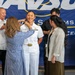 A U.S. Navy officer in white dress uniform is being pinned with new insignia during a promotion or commissioning ceremony. Family members surround her, with one person actively pinning her collar while others look on smiling. The event takes place in front of a Navy banner reading 'Naval... Systems Command... Center.'