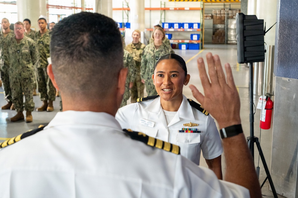 A military ceremony appears to be taking place inside a warehouse or hangar facility, likely a promotion or re-enlistment oath ceremony. A U.S. Navy officer in white dress uniform is raising her right hand while facing another officer whose back is to the camera. A formation of sailors and soldiers in camouflage uniforms observes in the background.