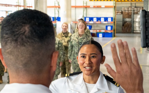 A military ceremony appears to be taking place inside a warehouse or hangar facility, likely a promotion or re-enlistment oath ceremony. A U.S. Navy officer in white dress uniform is raising her right hand while facing another officer whose back is to the camera. A formation of sailors and soldiers in camouflage uniforms observes in the background.