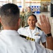 A military ceremony appears to be taking place inside a warehouse or hangar facility, likely a promotion or re-enlistment oath ceremony. A U.S. Navy officer in white dress uniform is raising her right hand while facing another officer whose back is to the camera. A formation of sailors and soldiers in camouflage uniforms observes in the background.