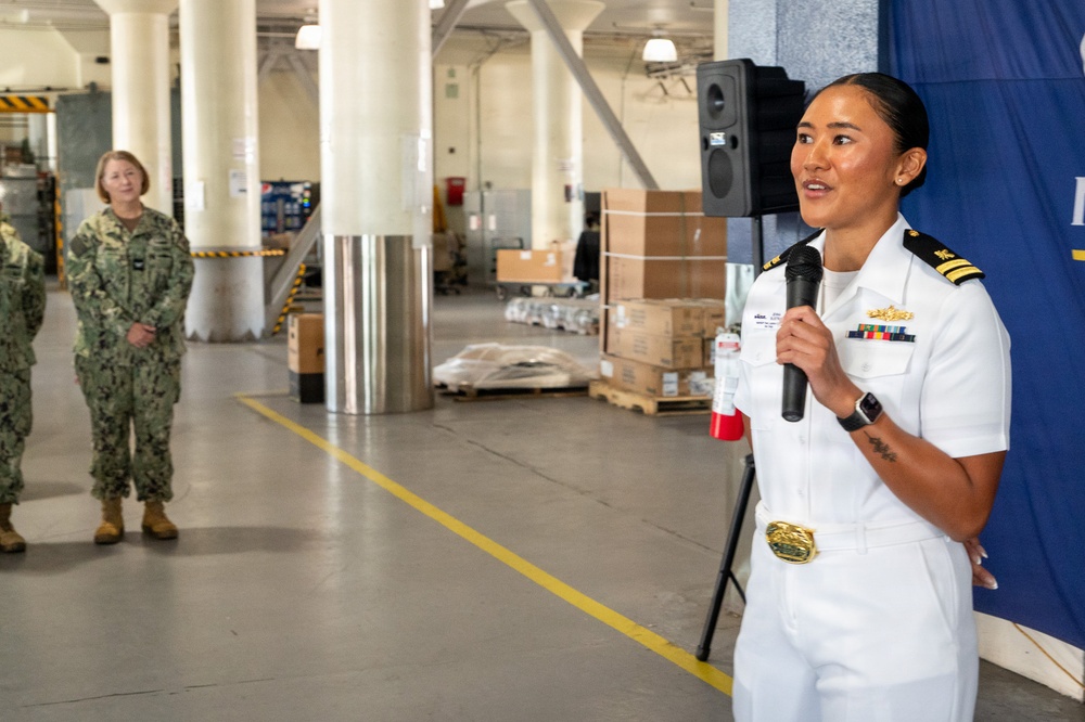 A U.S. Navy officer in white dress uniform is speaking into a microphone at what appears to be an indoor military facility or hangar. She is wearing rank insignia, service ribbons, and a gold belt buckle, suggesting a formal or ceremonial event. Two other personnel in camouflage uniforms stand in the background observing.