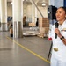 A U.S. Navy officer in white dress uniform is speaking into a microphone at what appears to be an indoor military facility or hangar. She is wearing rank insignia, service ribbons, and a gold belt buckle, suggesting a formal or ceremonial event. Two other personnel in camouflage uniforms stand in the background observing.