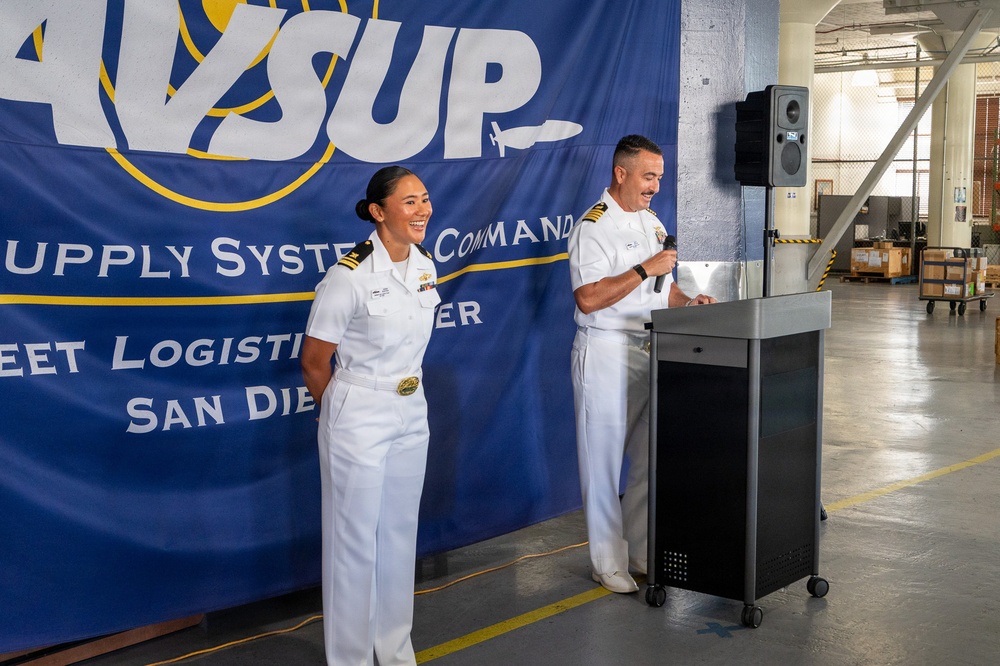 Two U.S. Navy officers in white dress uniforms stand before a podium at an official event for Naval Supply Systems Command (NAVSUP) Fleet Logistics Center San Diego. One officer holds a microphone at the podium while the other stands alongside, both appearing engaged and smiling. The event appears to be taking place inside a warehouse or logistics facility.