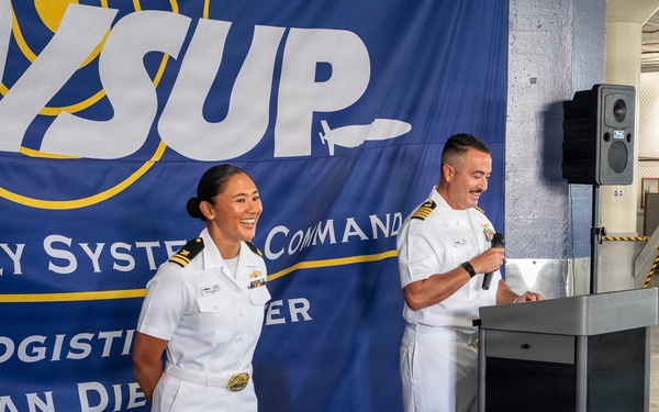 Two U.S. Navy officers in white dress uniforms stand before a podium at an official event for Naval Supply Systems Command (NAVSUP) Fleet Logistics Center San Diego. One officer holds a microphone at the podium while the other stands alongside, both appearing engaged and smiling. The event appears to be taking place inside a warehouse or logistics facility.