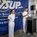 Two U.S. Navy officers in white dress uniforms stand before a podium at an official event for Naval Supply Systems Command (NAVSUP) Fleet Logistics Center San Diego. One officer holds a microphone at the podium while the other stands alongside, both appearing engaged and smiling. The event appears to be taking place inside a warehouse or logistics facility.