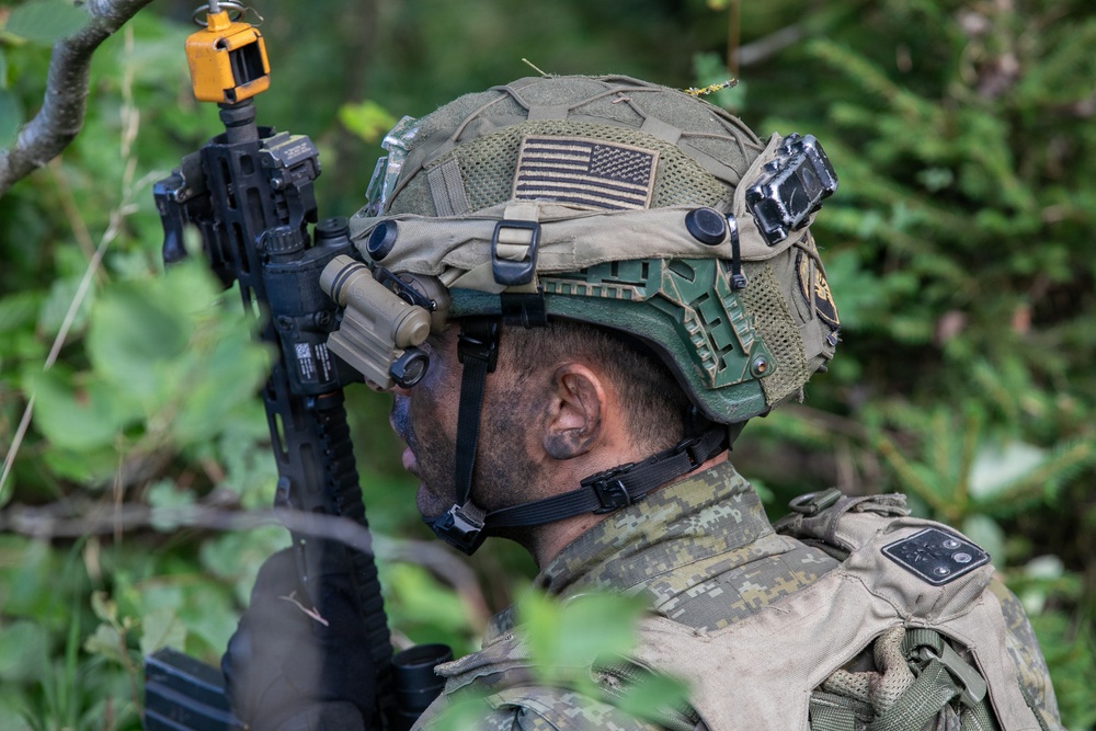 A soldier is photographed from behind and to the side in a wooded environment, equipped with advanced tactical gear. The individual wears a modern combat helmet adorned with a subdued U.S. flag patch and multiple mounted accessories. They are holding an assault rifle with attachments while appearing to observe their surroundings.