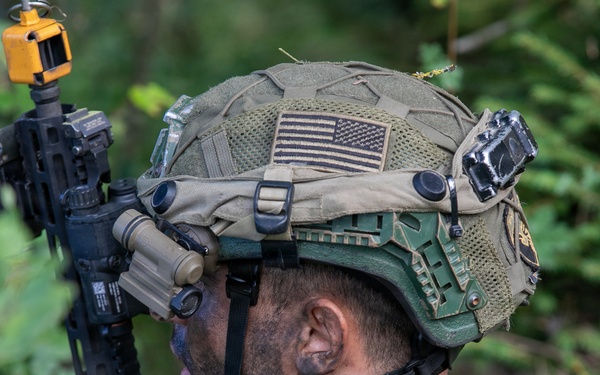 A soldier is photographed from behind and to the side in a wooded environment, equipped with advanced tactical gear. The individual wears a modern combat helmet adorned with a subdued U.S. flag patch and multiple mounted accessories. They are holding an assault rifle with attachments while appearing to observe their surroundings.