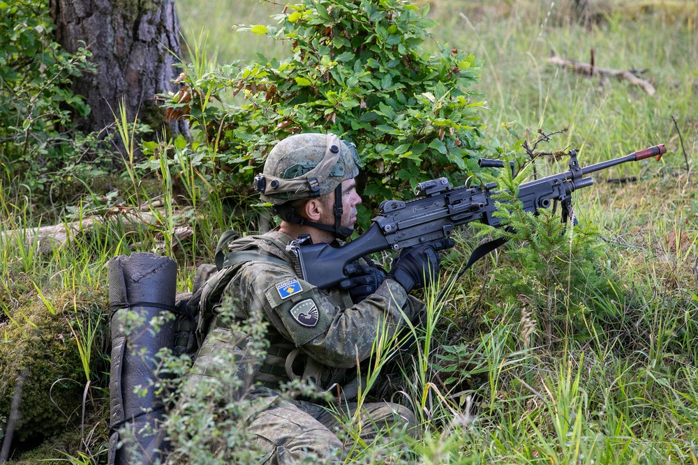A soldier in camouflage uniform is positioned in a prone/low crouch stance in a wooded, grassy area, aiming a light machine gun forward. The soldier wears a Kosovo Security Force (KSF) patch on the left shoulder, identifiable by the Kosovo flag insignia. The setting appears to be a field training exercise based on the environment and equipment.