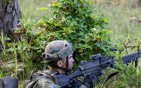 A soldier in camouflage uniform is positioned in a prone/low crouch stance in a wooded, grassy area, aiming a light machine gun forward. The soldier wears a Kosovo Security Force (KSF) patch on the left shoulder, identifiable by the Kosovo flag insignia. The setting appears to be a field training exercise based on the environment and equipment.