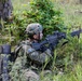 A soldier in camouflage uniform is positioned in a prone/low crouch stance in a wooded, grassy area, aiming a light machine gun forward. The soldier wears a Kosovo Security Force (KSF) patch on the left shoulder, identifiable by the Kosovo flag insignia. The setting appears to be a field training exercise based on the environment and equipment.