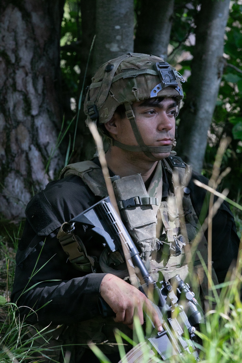 A young male soldier is positioned in a wooded area, seated among tall grass and trees. He is wearing full tactical gear including a combat helmet with a night-vision mount bracket, a plate carrier vest with magazine pouches, and is holding a scoped rifle. His expression is alert and focused, scanning to his left.