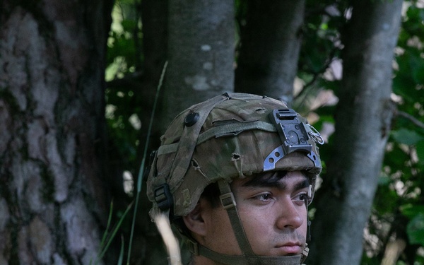 A young male soldier is positioned in a wooded area, seated among tall grass and trees. He is wearing full tactical gear including a combat helmet with a night-vision mount bracket, a plate carrier vest with magazine pouches, and is holding a scoped rifle. His expression is alert and focused, scanning to his left.