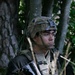 A young male soldier is positioned in a wooded area, seated among tall grass and trees. He is wearing full tactical gear including a combat helmet with a night-vision mount bracket, a plate carrier vest with magazine pouches, and is holding a scoped rifle. His expression is alert and focused, scanning to his left.