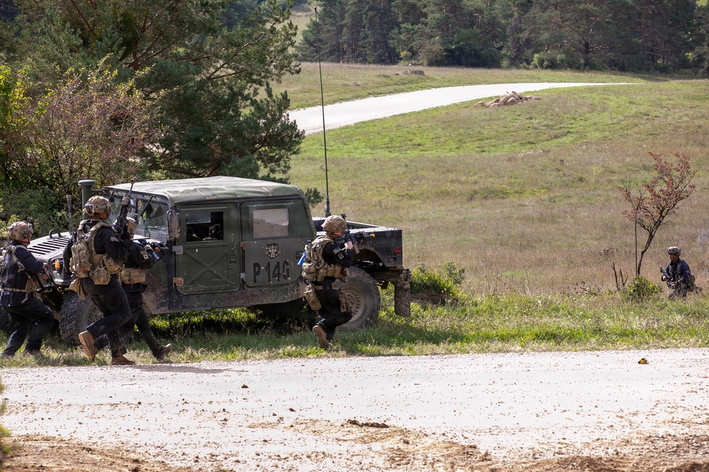The image shows military personnel in tactical gear conducting what appears to be a training exercise near a military HMMWV (Humvee) marked 'P-145'. Soldiers dressed in black uniforms with tan/coyote tactical vests are moving in formation around the vehicle, with at least one soldier aiming a weapon. The setting is an open field or training range with a paved road visible in the background.