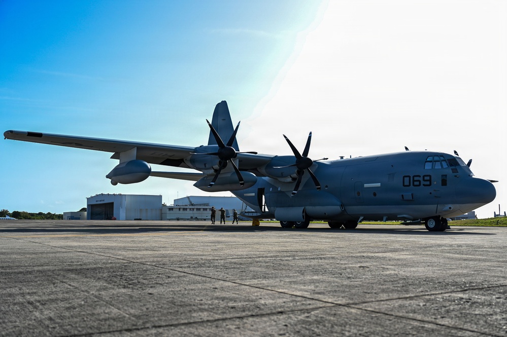 A large gray military turboprop transport aircraft marked '069' is parked on a concrete tarmac at a military airfield. The aircraft appears to be a C-130J Super Hercules variant, identifiable by its six-bladed propellers and gray livery consistent with U.S. Marine Corps markings. Several ground crew personnel are visible near the aircraft, with hangar buildings visible in the background.