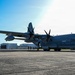 A large gray military turboprop transport aircraft marked '069' is parked on a concrete tarmac at a military airfield. The aircraft appears to be a C-130J Super Hercules variant, identifiable by its six-bladed propellers and gray livery consistent with U.S. Marine Corps markings. Several ground crew personnel are visible near the aircraft, with hangar buildings visible in the background.