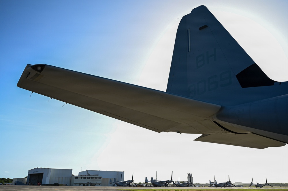 A low-angle ground-level photograph captures the underside of a large military transport aircraft's wing and tail section on a flight line. The tail section displays partial markings including the letters 'BH' and partial serial numbers. In the background, multiple tiltrotor aircraft (likely V-22 Ospreys) are parked on the ramp alongside military hangars and a control tower.