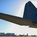A low-angle ground-level photograph captures the underside of a large military transport aircraft's wing and tail section on a flight line. The tail section displays partial markings including the letters 'BH' and partial serial numbers. In the background, multiple tiltrotor aircraft (likely V-22 Ospreys) are parked on the ramp alongside military hangars and a control tower.