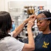 A technician wearing a face mask and glasses is measuring the head circumference of a woman wearing a navy blue military-style shirt with an eagle/anchor insignia. The measurement is being taken using a flexible tape measure across the woman's forehead, likely for fitting a helmet or protective headgear. A third person is visible in the blurred background of what appears to be a supply or equipment fitting room.