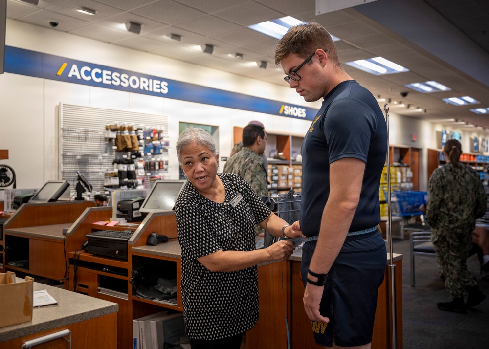 A store employee is measuring the waist of a young man wearing a Navy physical training uniform inside what appears to be a military uniform/exchange store. The employee, an older woman wearing a black patterned blouse with a name tag, is using a measuring tape around the customer's midsection. The store background shows sections labeled 'Accessories' and 'Shoes' with other military personnel visible in the background.