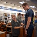 A store employee is measuring the waist of a young man wearing a Navy physical training uniform inside what appears to be a military uniform/exchange store. The employee, an older woman wearing a black patterned blouse with a name tag, is using a measuring tape around the customer's midsection. The store background shows sections labeled 'Accessories' and 'Shoes' with other military personnel visible in the background.