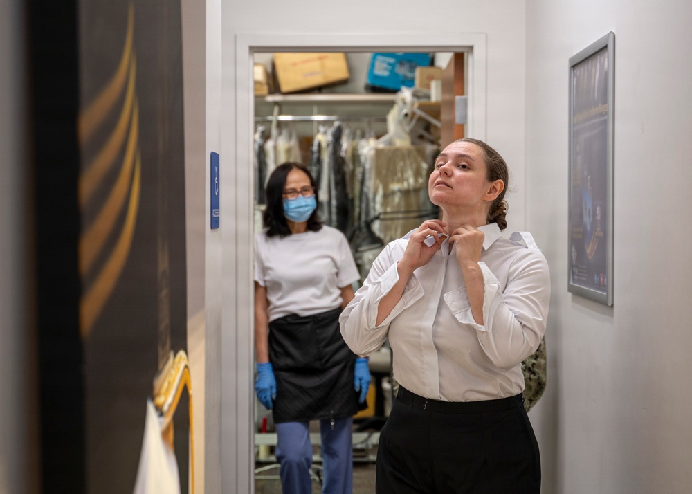 A woman in a white dress shirt and black skirt adjusts her collar or neckline, appearing to be getting dressed or fitted. In the background, another woman wearing a face mask, black apron, and blue gloves stands in a doorway leading to what appears to be a dry cleaning or tailoring storage room with garments on hangers. The setting suggests a uniform fitting or tailoring service environment.