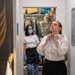A woman in a white dress shirt and black skirt adjusts her collar or neckline, appearing to be getting dressed or fitted. In the background, another woman wearing a face mask, black apron, and blue gloves stands in a doorway leading to what appears to be a dry cleaning or tailoring storage room with garments on hangers. The setting suggests a uniform fitting or tailoring service environment.