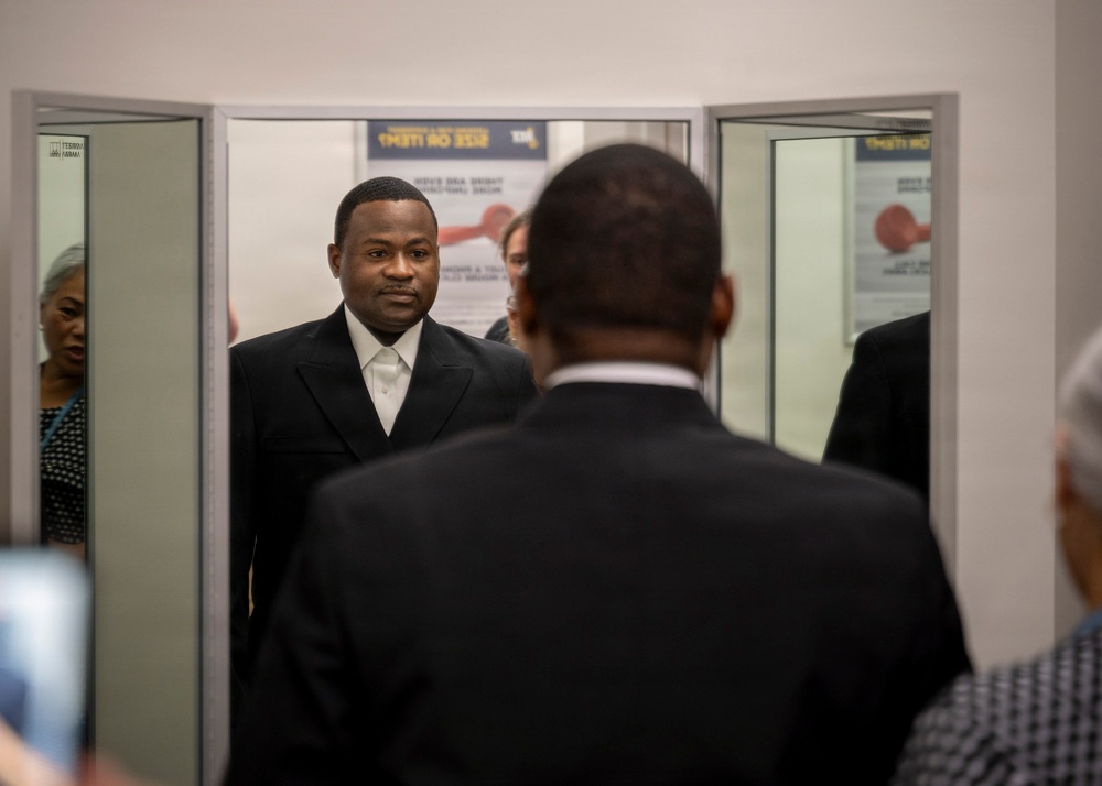 The image shows several individuals in formal dark attire gathered near a set of glass doors or a security checkpoint area. A man in a black suit with a white shirt faces the camera while another individual stands with their back to the viewer in the foreground. Mirrored or glass panels on either side create reflections of additional people in the space.