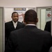The image shows several individuals in formal dark attire gathered near a set of glass doors or a security checkpoint area. A man in a black suit with a white shirt faces the camera while another individual stands with their back to the viewer in the foreground. Mirrored or glass panels on either side create reflections of additional people in the space.