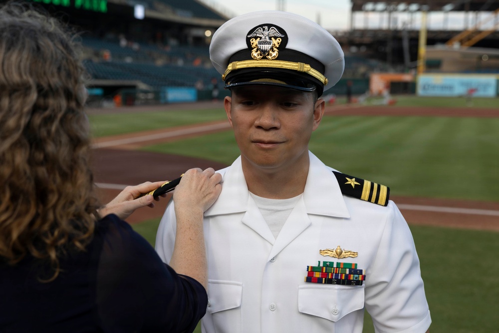 A U.S. Navy officer in white dress uniform is having insignia or rank devices pinned to his shoulder by a woman at what appears to be a baseball stadium. The officer wears a white combination cover with gold insignia, two gold stripes with a star on his shoulder boards indicating the rank of Lieutenant Commander, and several service ribbons with a warfare device on his chest. This appears to be a military promotion or commissioning ceremony conducted at a professional baseball venue.