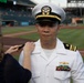A U.S. Navy officer in white dress uniform is having insignia or rank devices pinned to his shoulder by a woman at what appears to be a baseball stadium. The officer wears a white combination cover with gold insignia, two gold stripes with a star on his shoulder boards indicating the rank of Lieutenant Commander, and several service ribbons with a warfare device on his chest. This appears to be a military promotion or commissioning ceremony conducted at a professional baseball venue.