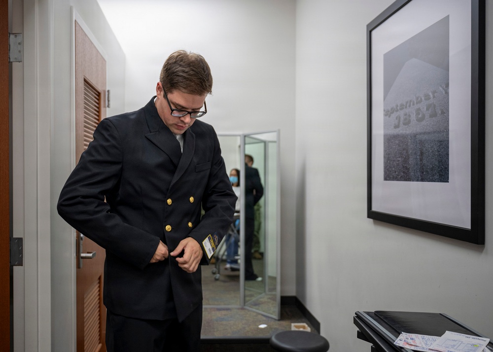 A young man wearing glasses and a dark navy double-breasted jacket with gold buttons is adjusting or buttoning his jacket in what appears to be a hallway or anteroom. A framed black-and-white photograph is visible on the wall to his right, and a mirror in the background reflects another person wearing a face mask. The setting appears to be an office or institutional building.
