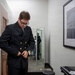 A young man wearing glasses and a dark navy double-breasted jacket with gold buttons is adjusting or buttoning his jacket in what appears to be a hallway or anteroom. A framed black-and-white photograph is visible on the wall to his right, and a mirror in the background reflects another person wearing a face mask. The setting appears to be an office or institutional building.