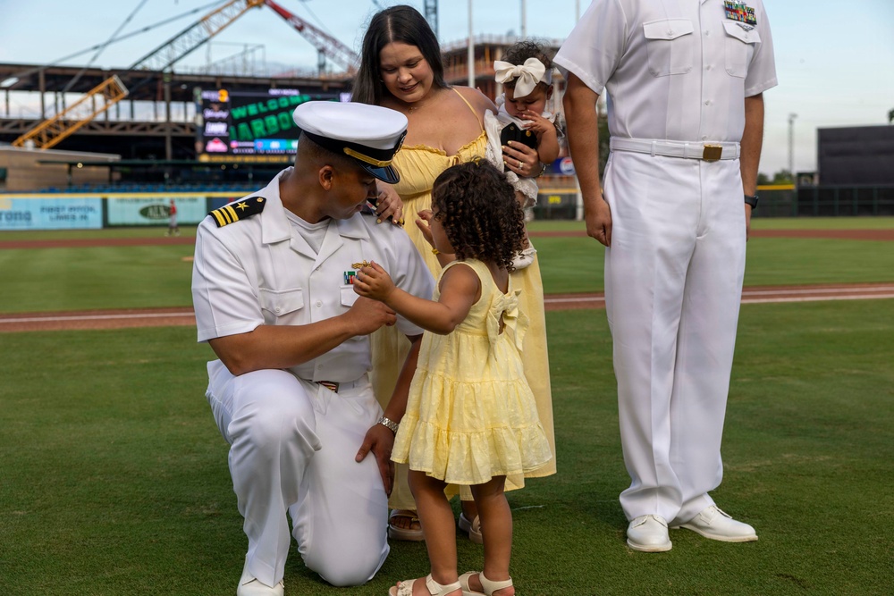 A U.S. Navy officer in white dress uniform kneels on a baseball field while a young girl in a yellow dress appears to pin something onto his uniform, likely during a promotion or award ceremony. A woman in a yellow dress holds a baby in the background, and a second Navy officer stands nearby. The event appears to be taking place at a minor league baseball stadium during a military appreciation event.