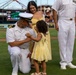 A U.S. Navy officer in white dress uniform kneels on a baseball field while a young girl in a yellow dress appears to pin something onto his uniform, likely during a promotion or award ceremony. A woman in a yellow dress holds a baby in the background, and a second Navy officer stands nearby. The event appears to be taking place at a minor league baseball stadium during a military appreciation event.
