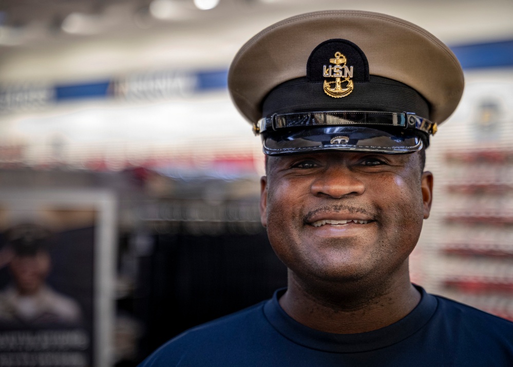 A smiling man wearing a United States Navy Chief Petty Officer combination cover (cap) with the USN anchor insignia in gold. He is dressed in a navy blue t-shirt and appears to be indoors, likely in a retail or exchange store environment. The portrait is a close-up shot with a blurred background.