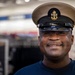 A smiling man wearing a United States Navy Chief Petty Officer combination cover (cap) with the USN anchor insignia in gold. He is dressed in a navy blue t-shirt and appears to be indoors, likely in a retail or exchange store environment. The portrait is a close-up shot with a blurred background.