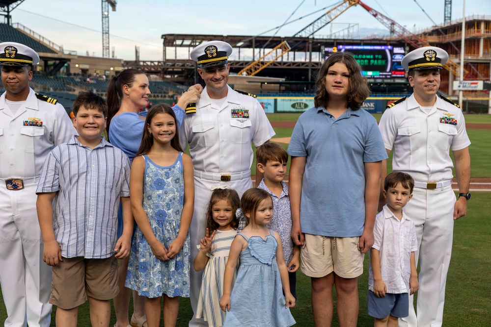 A group photo taken on a baseball field at what appears to be 'Armor Park' stadium, featuring two U.S. Navy officers in white dress uniforms alongside a woman and several children of varying ages. The setting suggests a military appreciation event or ceremony at a minor league baseball game. Construction cranes and stadium infrastructure are visible in the background.