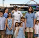 A group photo taken on a baseball field at what appears to be 'Armor Park' stadium, featuring two U.S. Navy officers in white dress uniforms alongside a woman and several children of varying ages. The setting suggests a military appreciation event or ceremony at a minor league baseball game. Construction cranes and stadium infrastructure are visible in the background.